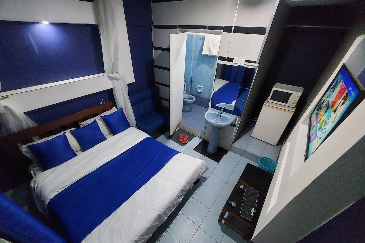 Quarto de hotel compacto com cama, uma pequena escrivaninha com computador, pia e espelho.