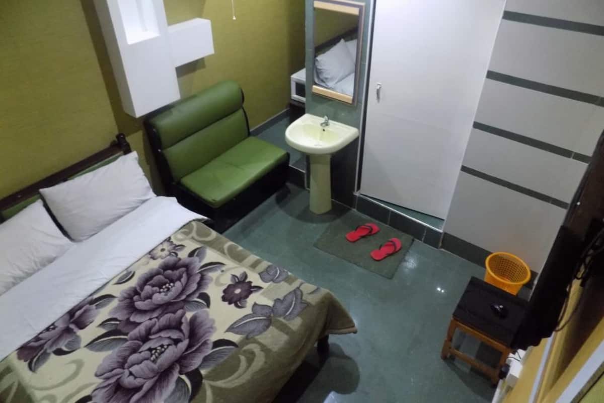 Quarto de hotel pequeno com cama, uma cadeira verde, pia, guarda-roupa e um par de chinelos vermelhos no chão.