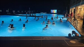 Une zone de piscine où se trouvent plusieurs personnes dans l’eau, un écran de télévision en arrière-plan et un bâtiment doté d’un espace extérieur aménagé pour s’asseoir.