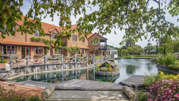A riverside view with a stone bench, brick buildings, and a small island with plants. - Monaco Resort - Home to Compass Cottage (Monaco)
