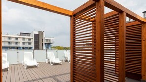 A wooden deck with white lounge chairs, a wooden pergola, and a view of modern buildings in the background. - Cozy Studio | 400 m to the Beach, Gym, Balcony (Kołobrzeg)