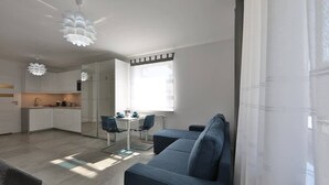 A modern living room with a sofa, a dining table with chairs, a kitchen area, and a large window with curtains. - Cozy Studio | 400 m to the Beach, Gym, Balcony (Kołobrzeg)