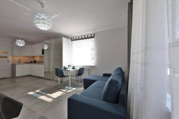 A modern living room with a sofa, a dining table with chairs, a kitchen area, and a large window with curtains.