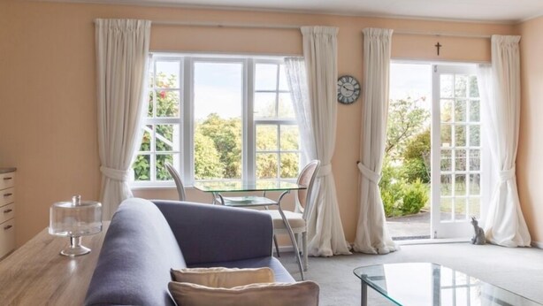 Ein gemütliches Zimmer mit einer Couch, einem Glastisch und Blick auf Grün hinter großen Fenstern.