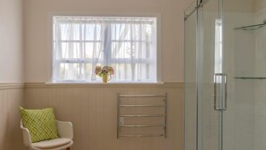 Ein Badezimmer mit einer Duschkabine aus Glas, einem weißen Stuhl mit gemustertem Sitzkissen, einem Fenster mit durchsichtigen Vorhängen und einem Handtuchhalter.