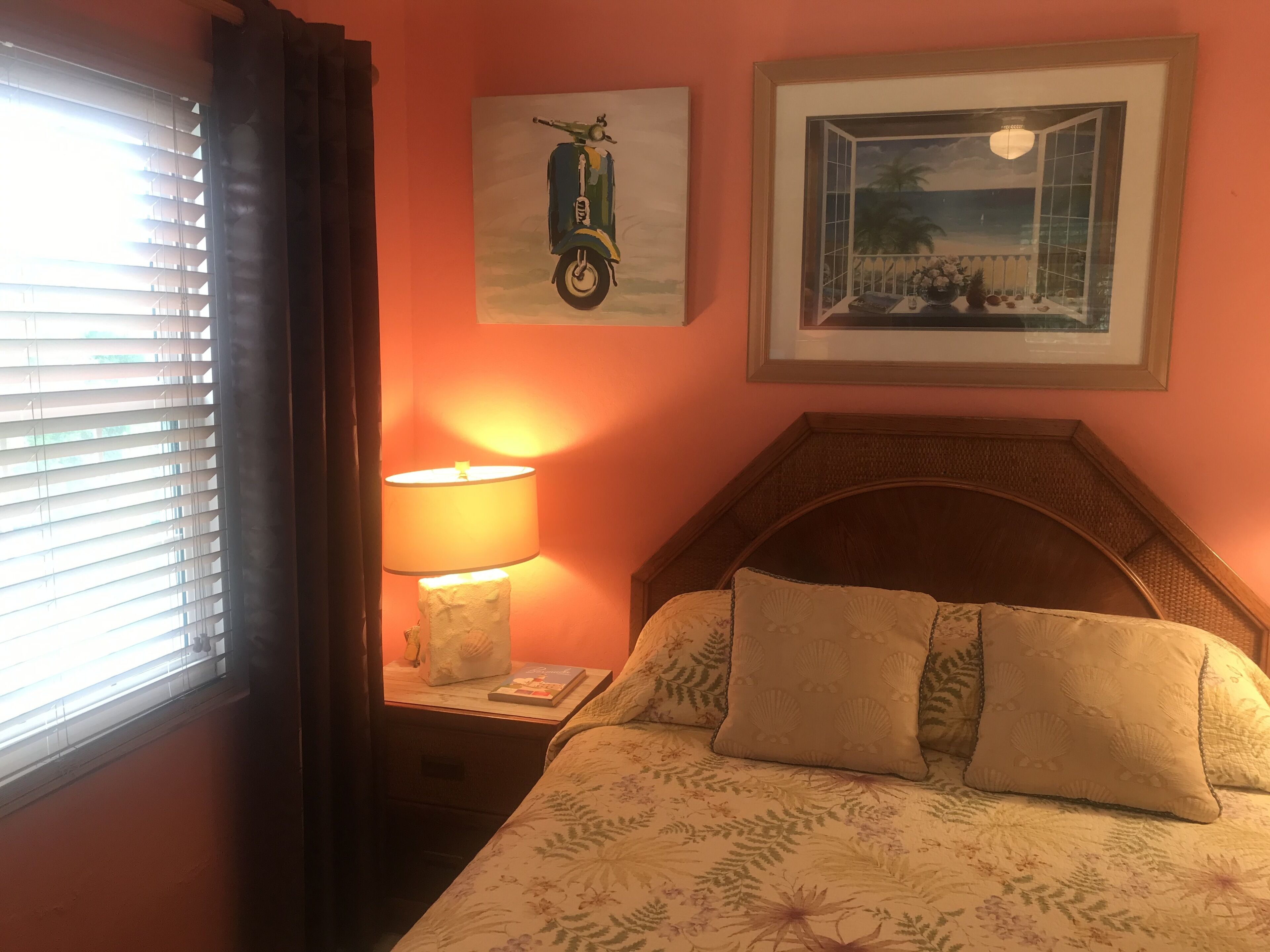 A bedroom with a bed, a nightstand with a lamp, a window with blinds, and two framed pictures on the wall.