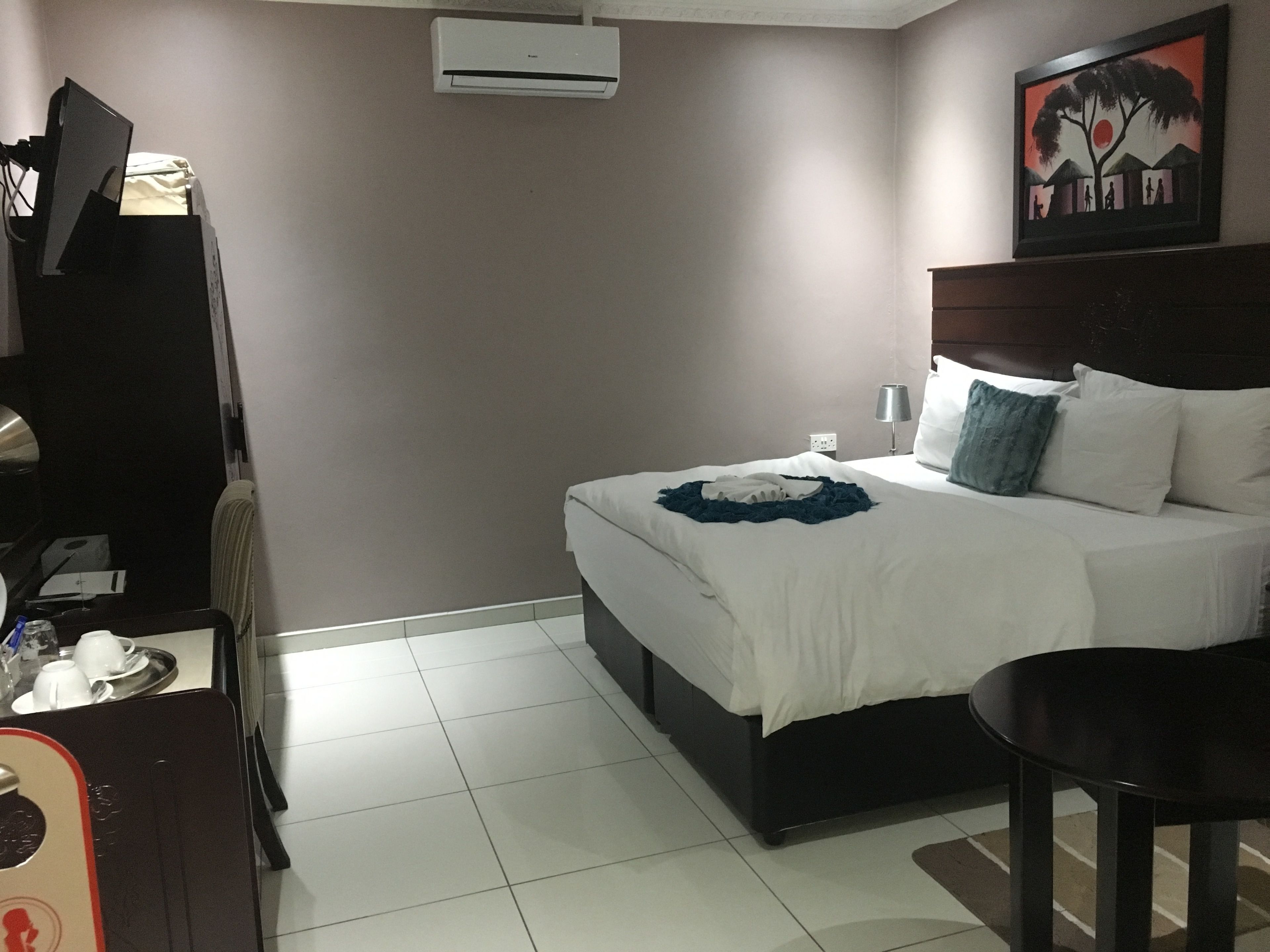A hotel room with a bed, bedside table, and a desk with a chair. There is an air conditioning unit on the wall and a painting above the bed.