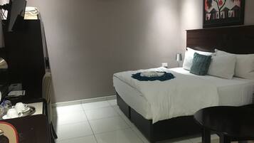 A hotel room with a bed, bedside table, and a desk with a chair. There is an air conditioning unit on the wall and a painting above the bed.