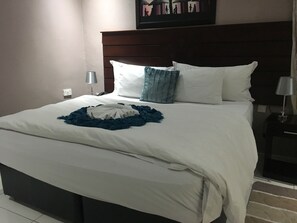 Una cama bien hecha con sábanas blancas, una almohada decorativa azul y una manta azul. Encima de la cama hay una obra de arte colgada en la pared y, a la derecha, una mesita de noche con una lámpara.