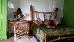 Une pièce avec une porte verte, un lit en bois avec des oreillers, une chaise en osier et un vase de fleurs.