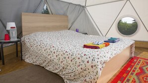 Una habitación tipo tienda de campaña con una cama, una mesita con lámpara y una ventana.