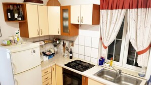 Eine kompakte Küche mit einem Gasherd, einem Geschirrspüler und einem Spülbecken. Ein Fenster mit Vorhängen und eine kleine Ablage über dem Herd, auf der Flaschen stehen.