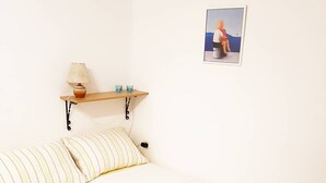 Ein schlichtes Zimmer mit einem Bett, einem kleinen Regal mit einer Lampe und einem Bild an der Wand.