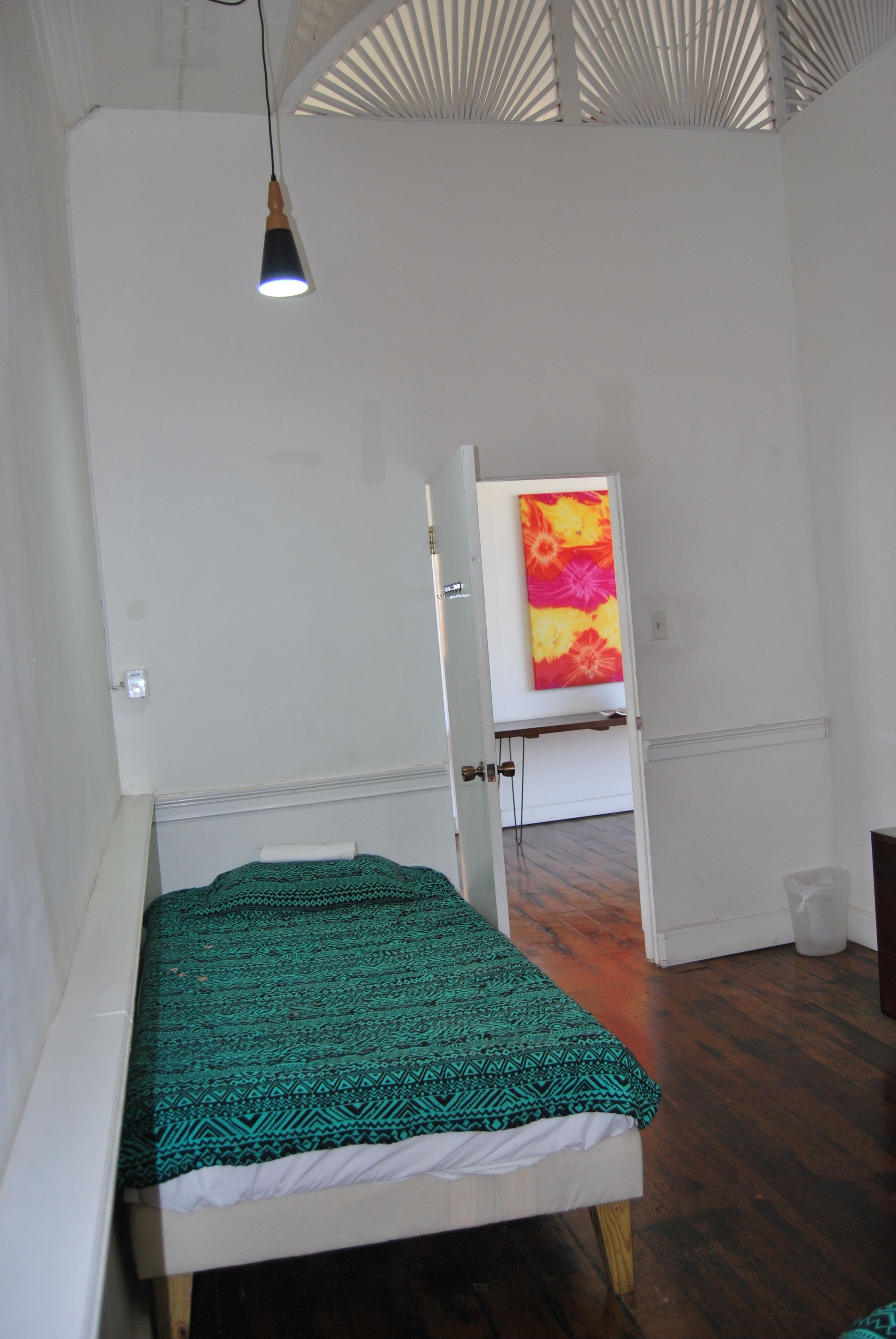 A room with a bed, a hanging light fixture, and a door leading to another room with a colorful painting.