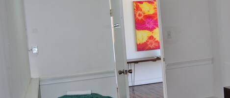 A room with a bed, a hanging light fixture, and a door leading to another room with a colorful painting.