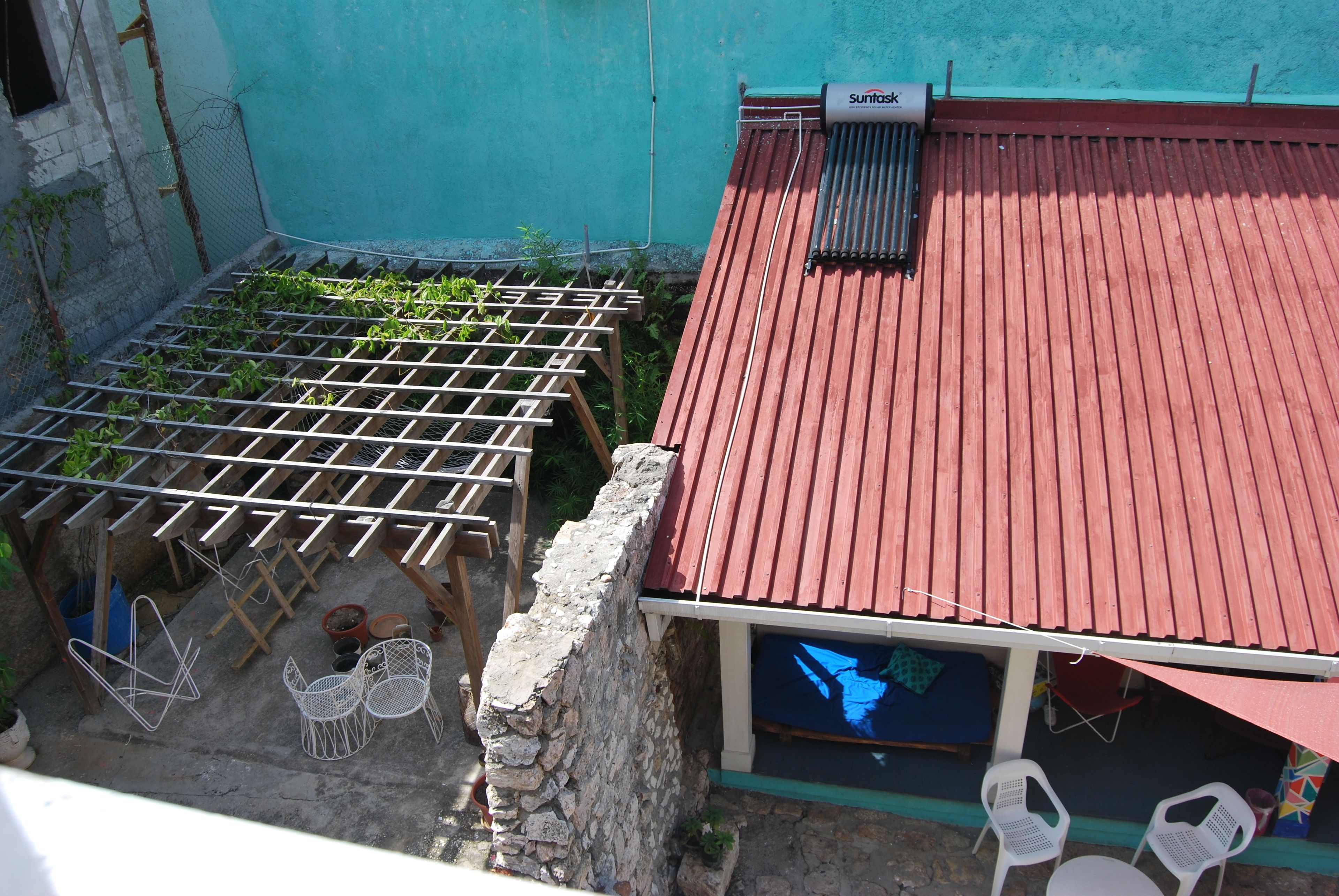 Un jardín en la azotea con techo de metal rojo, un muro de piedra y un espacio de descanso con sillas de plástico blanco.