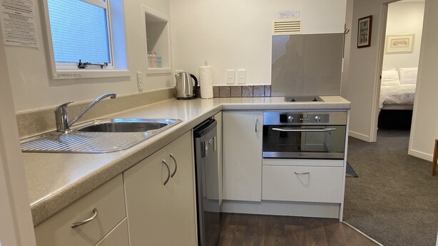 A compact kitchen with a sink, stove, and oven, a small window, and a door leading to a bedroom. - Heart of Russell Apartment - Russell Apartment (Russell)
