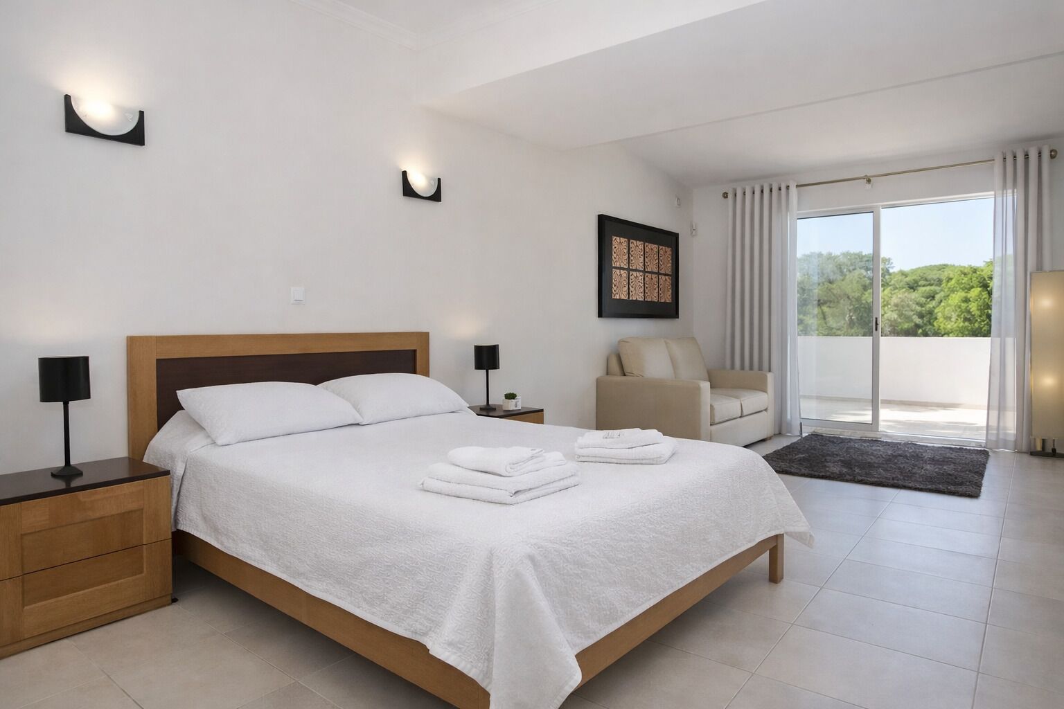 Ein modernes Schlafzimmer mit einem großen Bett, Nachttischen, einem Sessel und Blick ins Grüne durch das Fenster.