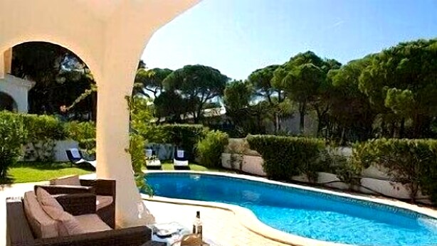 Una piscina de borde curvo, rodeada de vegetación y un patio con mobiliario exterior.