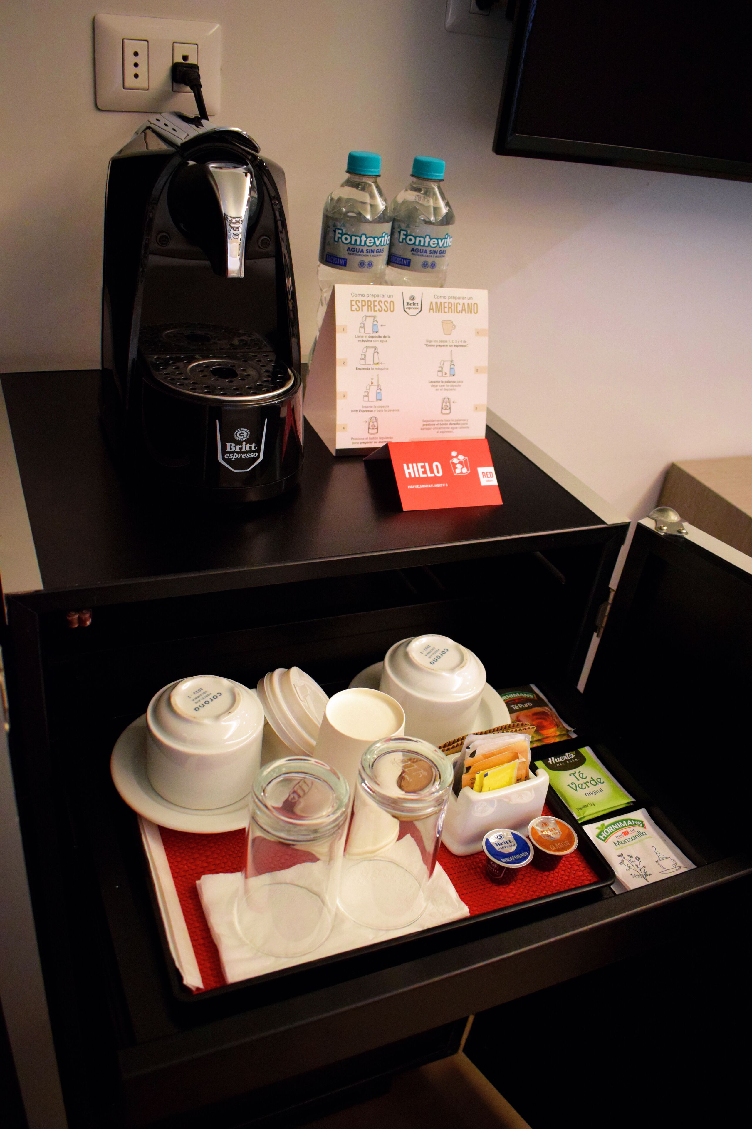 A hotel room's minibar with a coffee maker, water bottles, and various food items.