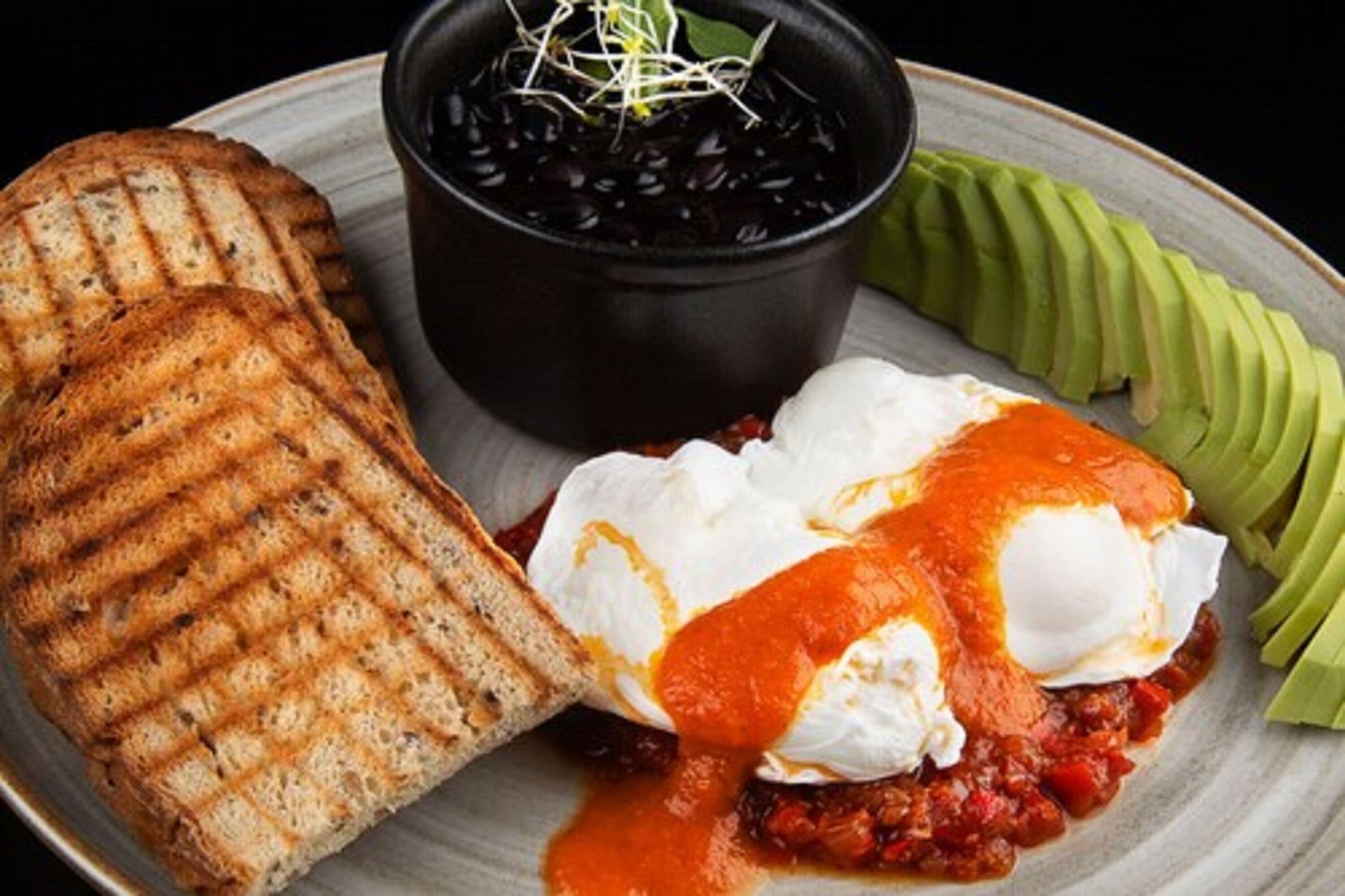 Un plato con tostadas a la parrilla, huevos escalfados, un dip de frijoles negros y aguacate en rodajas.