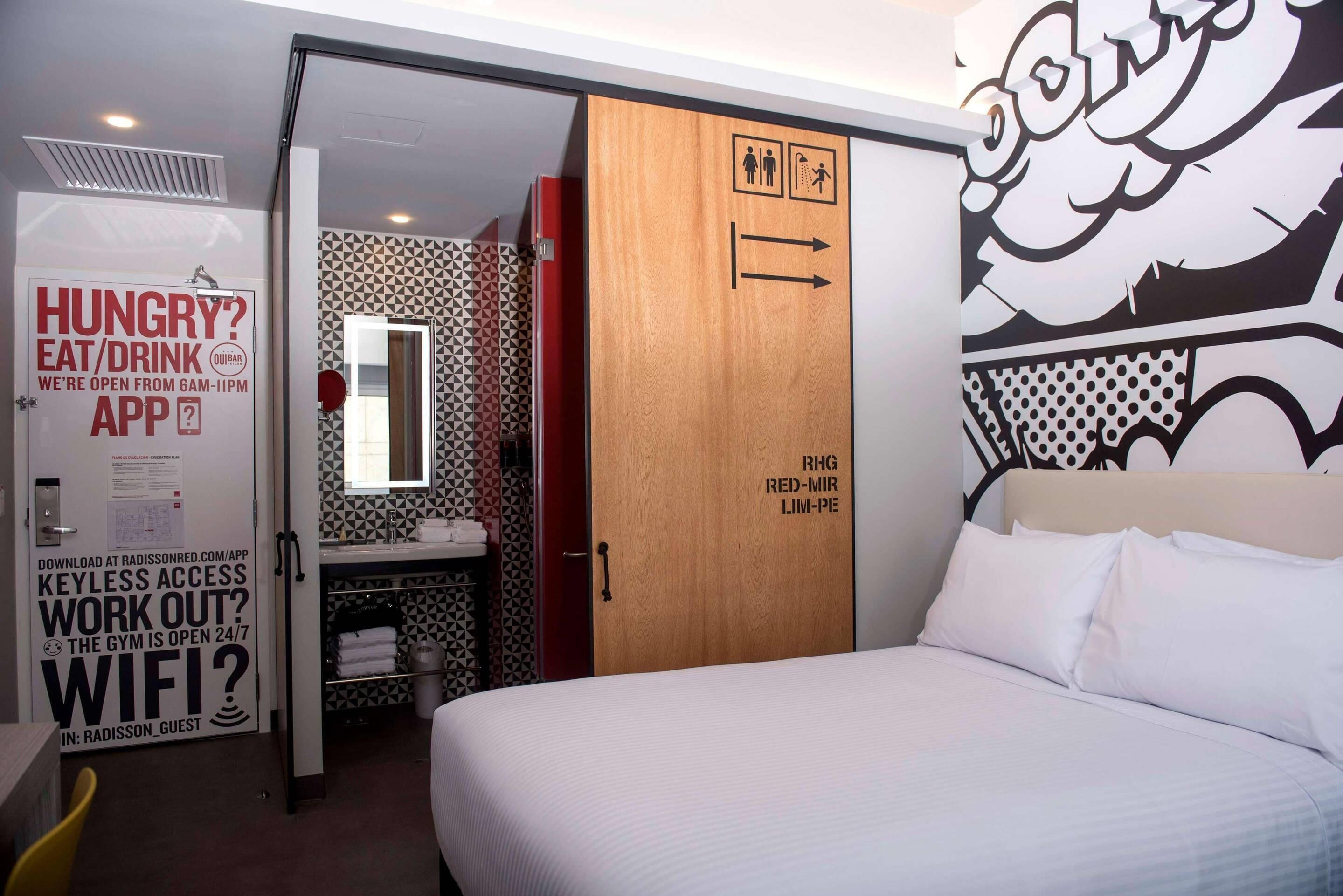 A hotel room with a bed, a bathroom with a mirror, and a wall with a geometric pattern.