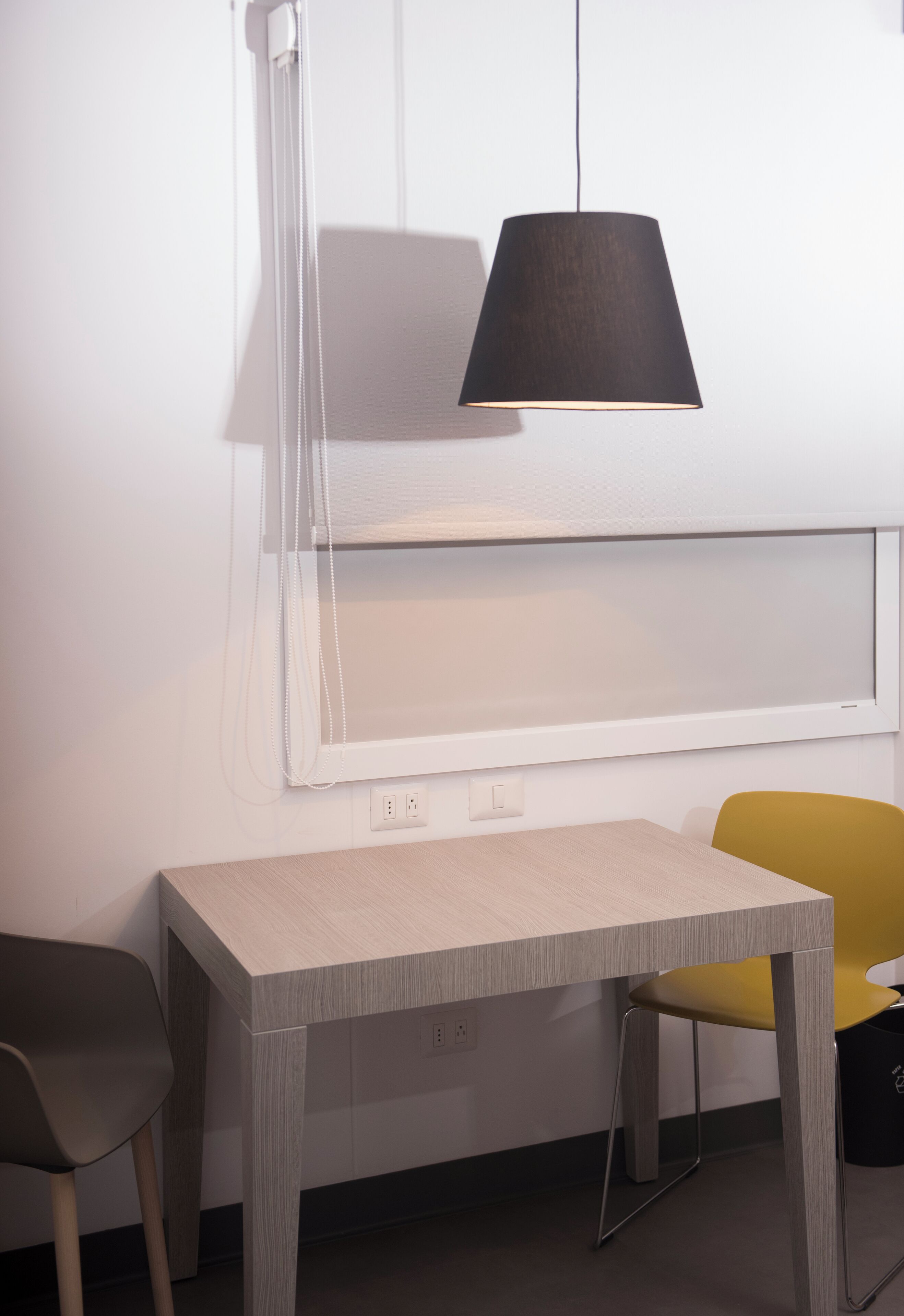 Una habitación moderna con un escritorio, dos sillas, una lámpara negra y un ventanal con persianas.