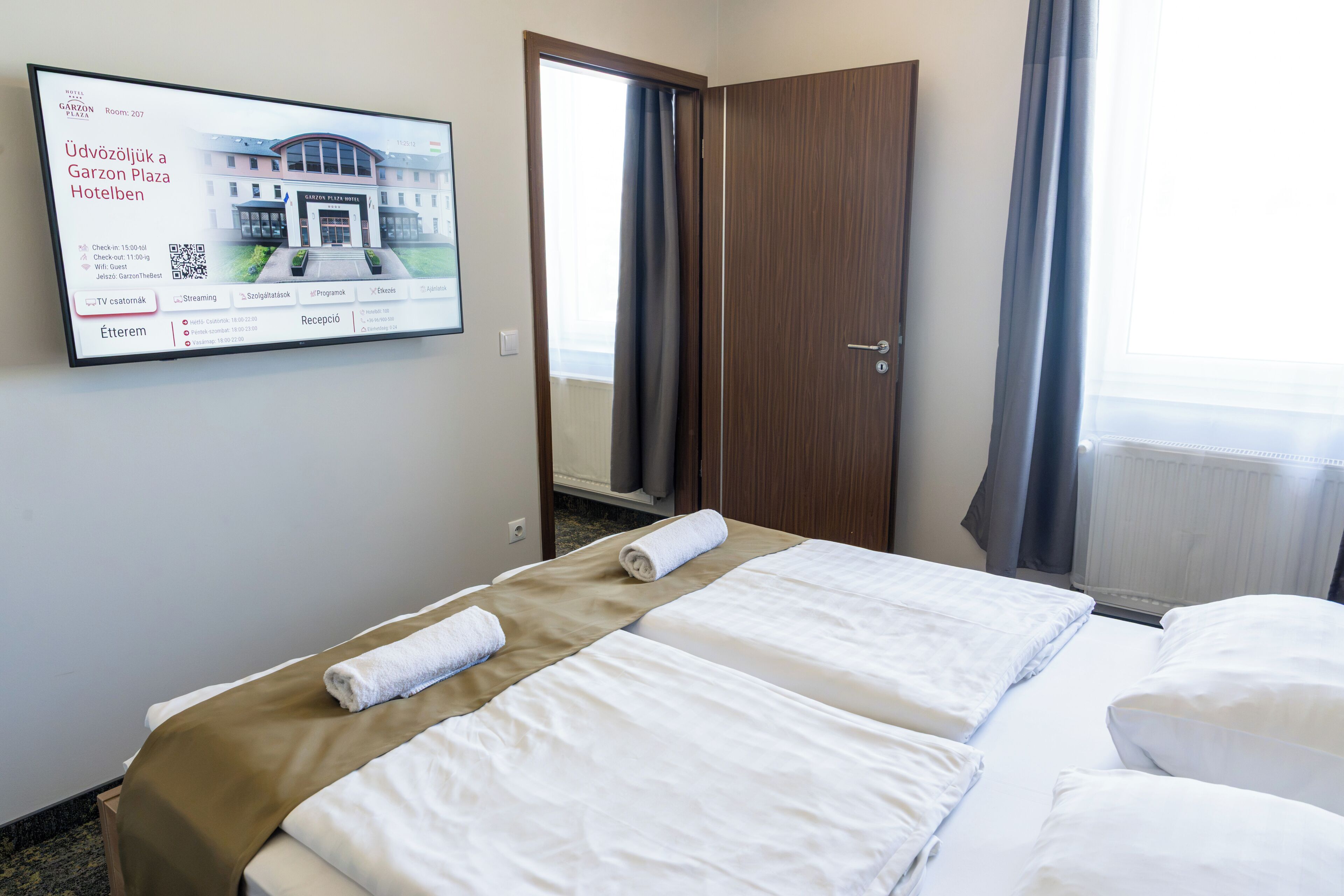 Une chambre d’hôtel avec deux lits, un écran de télévision affichant des informations sur l’hôtel et une porte donnant sur l’extérieur.