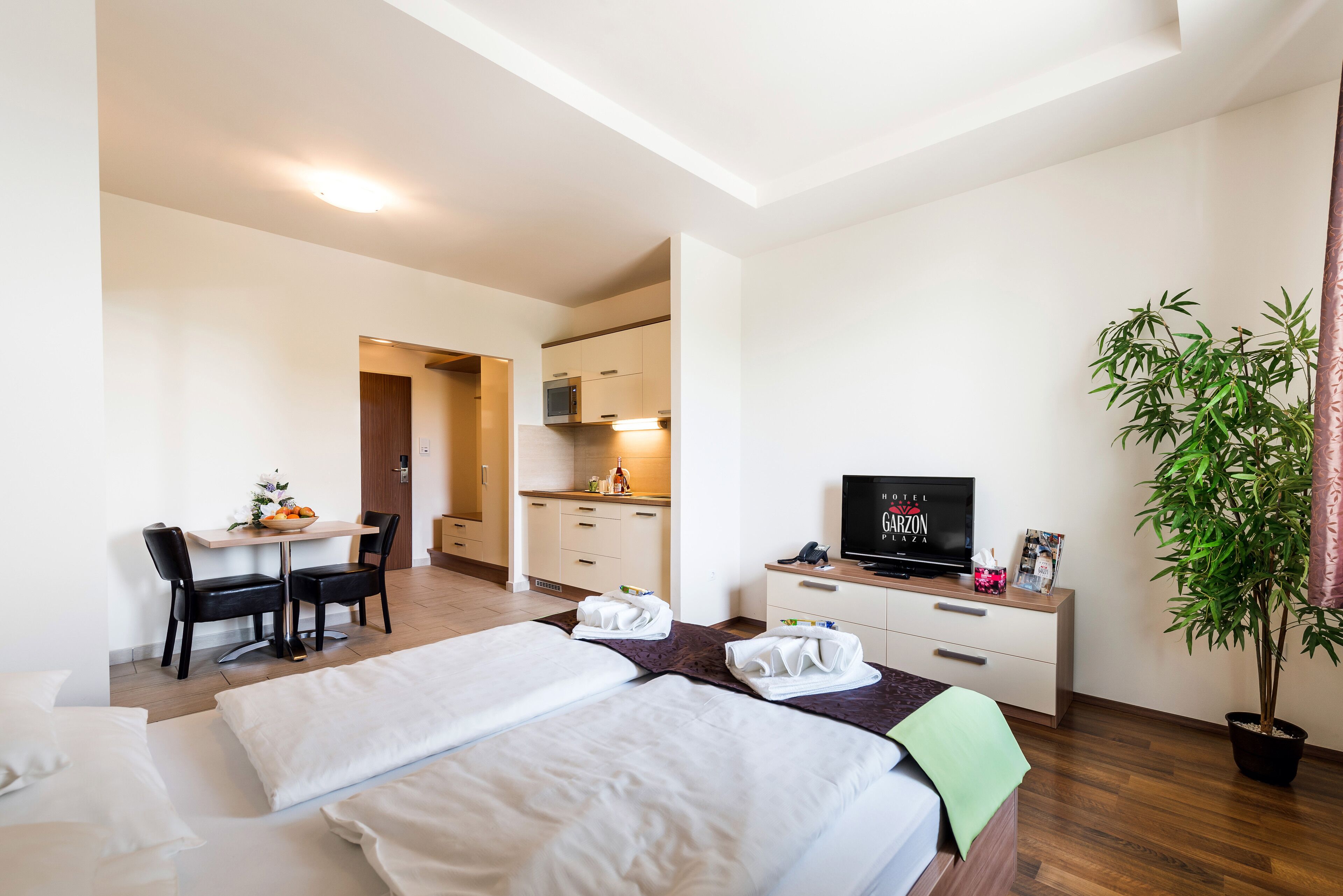 Ein Hotelzimmer mit zwei Betten, einem Schreibtisch mit Stuhl, einem Fernseher, einer Zimmerpflanze und einer Küchenzeile.