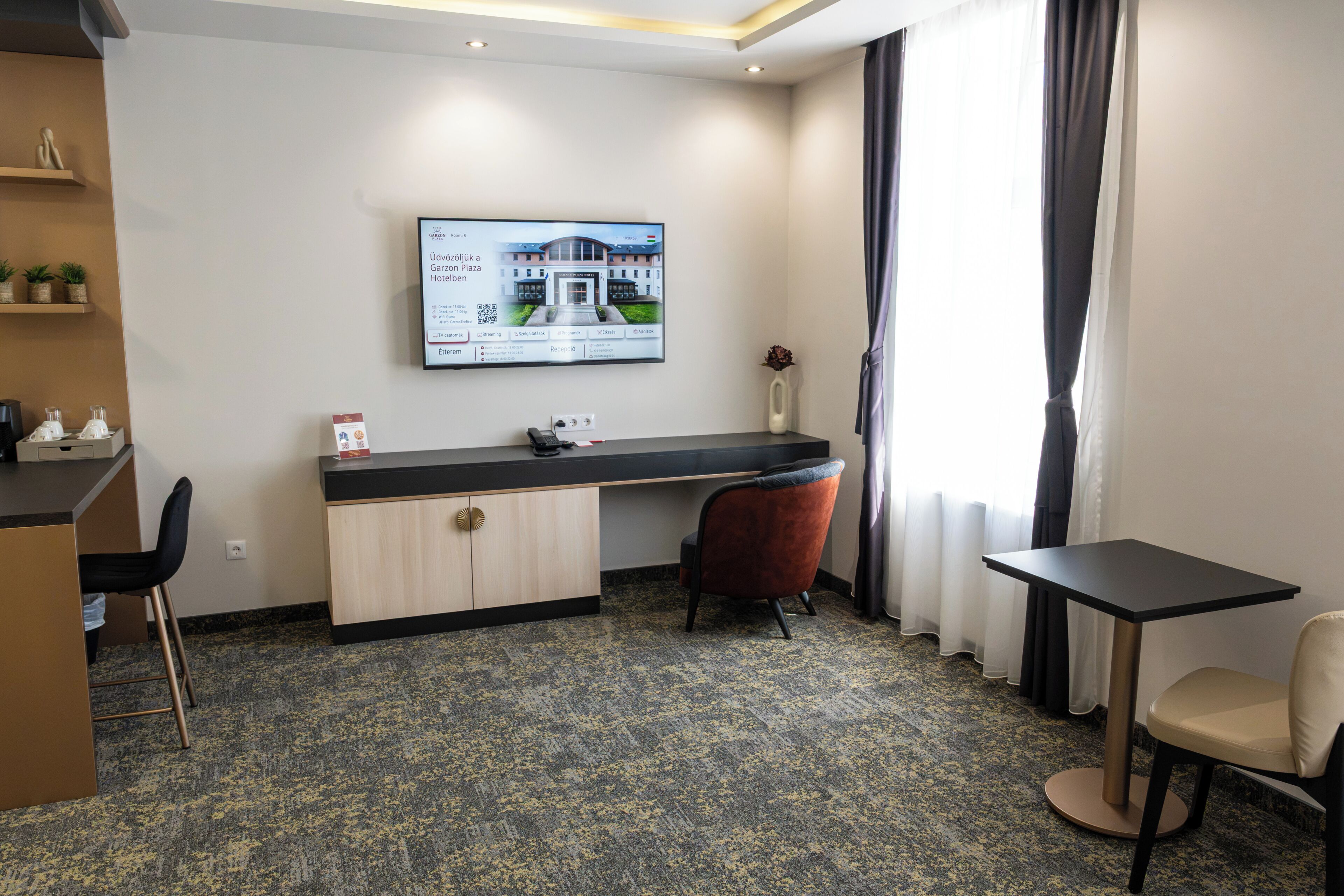 Ein Hotelzimmer mit Schreibtisch, zwei Stühlen, einem an der Wand montierten Fernseher und einem Fenster mit Vorhängen.