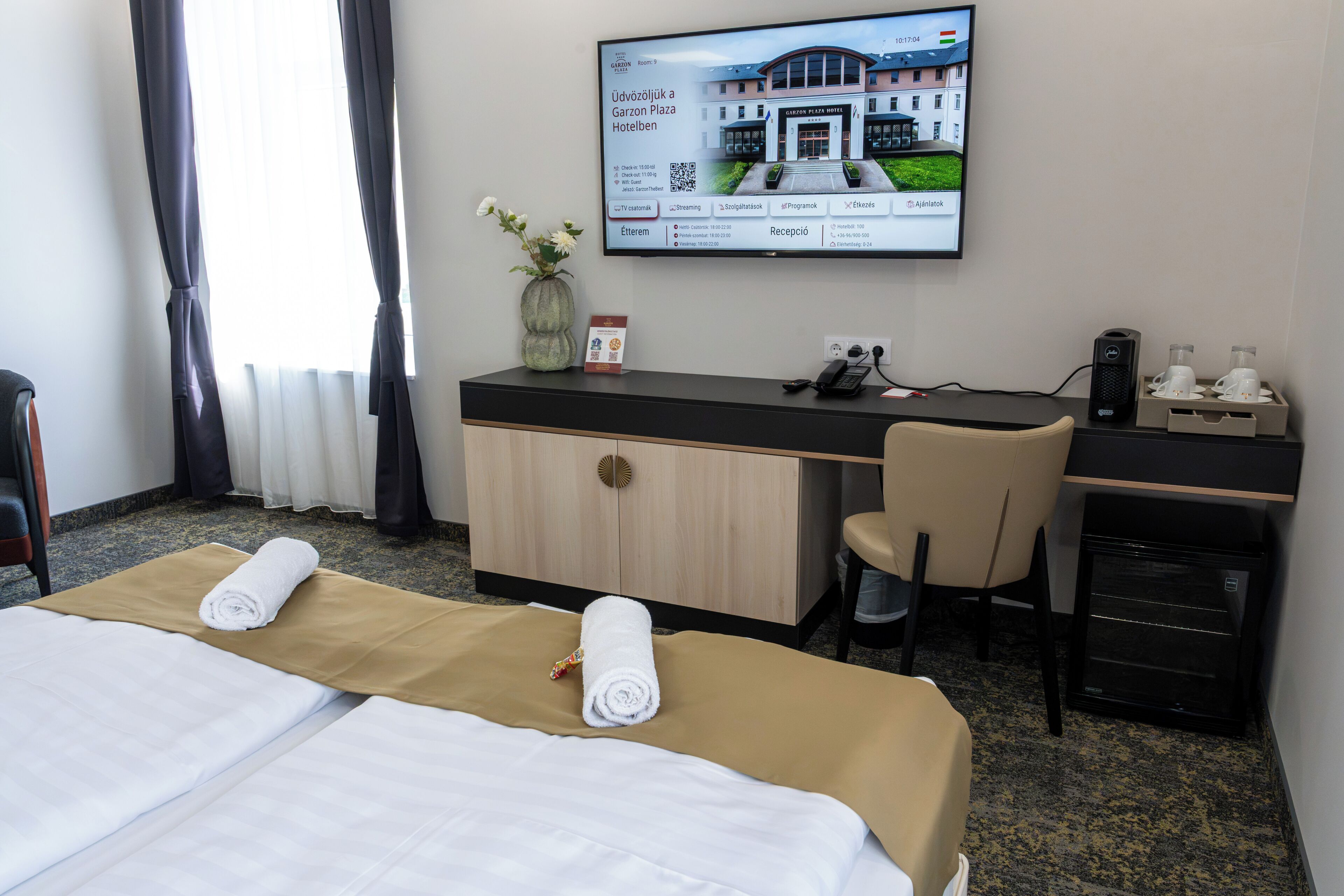 Habitación de hotel con cama, escritorio, silla, televisor mostrando una imagen de hotel y un refrigerador pequeño.