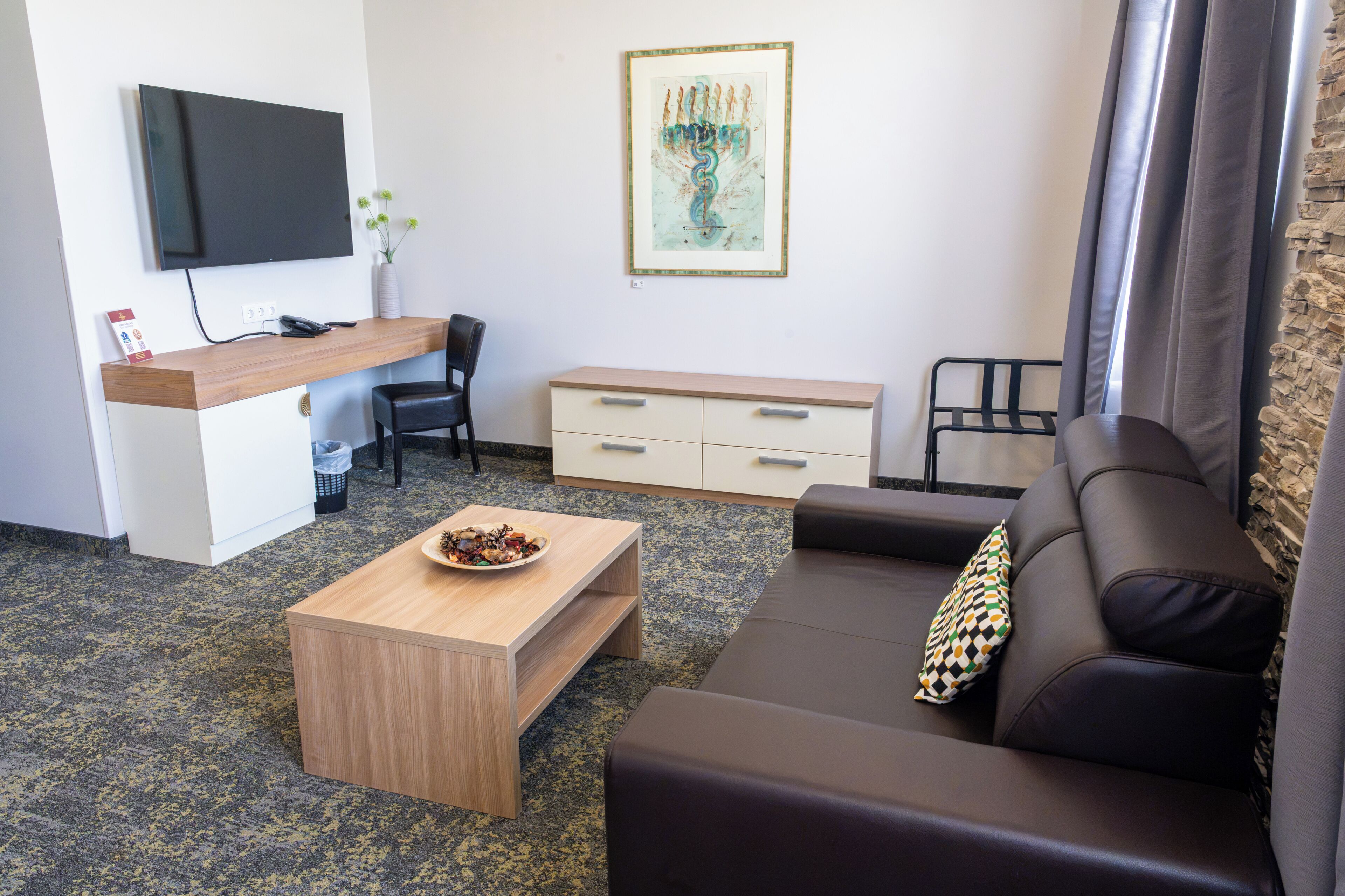 Una habitación moderna con un sofá, una mesita de centro de madera, un escritorio con una silla, un televisor montado en la pared y una obra de arte enmarcada.