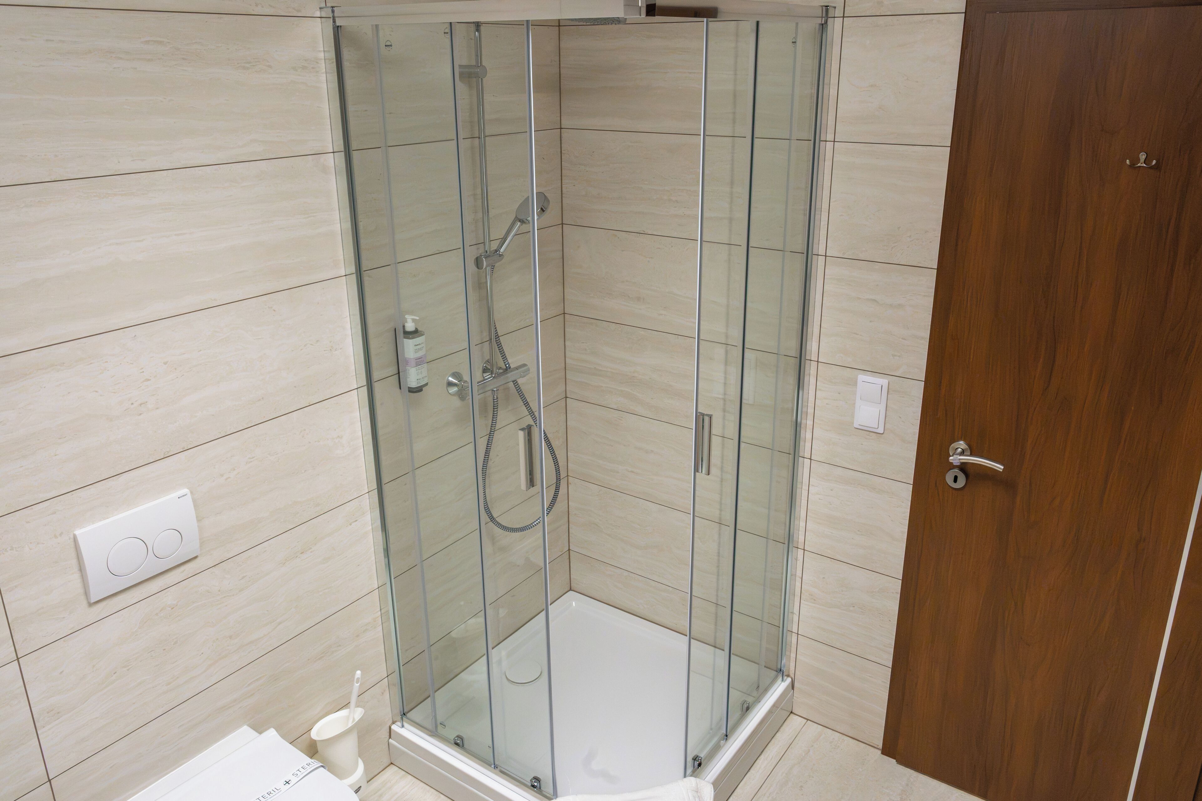 Eine Duschkabine mit Glastüren, eine Holztür, eine weiße Toilette und ein an der Wand befestigter Lichtschalter.