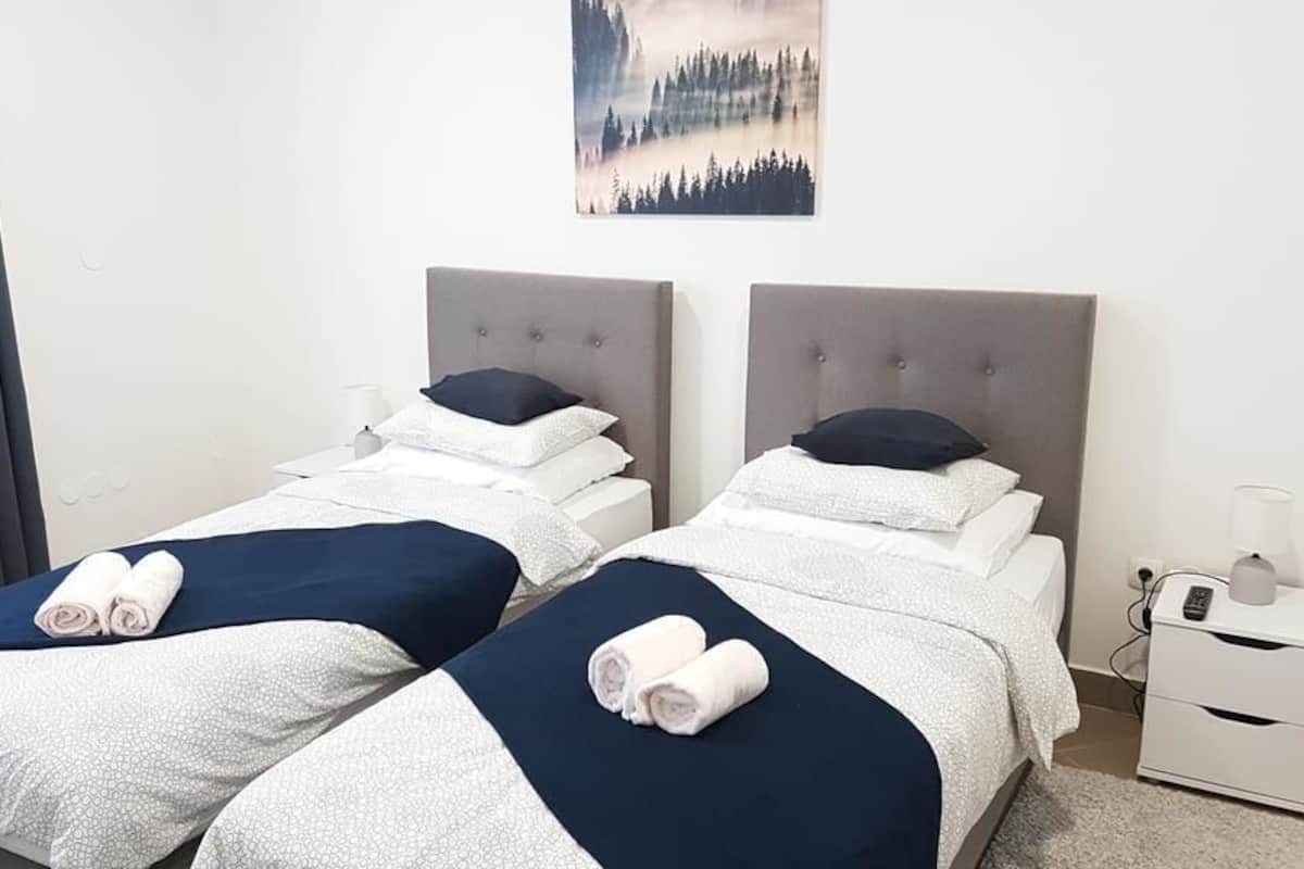 Ein Hotelzimmer mit zwei Betten, jeweils mit grauem Kopfteil, weißer Bettwäsche und dunkelblauen Akzenten. An der Wand über den Betten hängt ein Bild und neben jedem Bett steht ein kleiner Nachttisch mit einer Lampe und einer Ladestation.