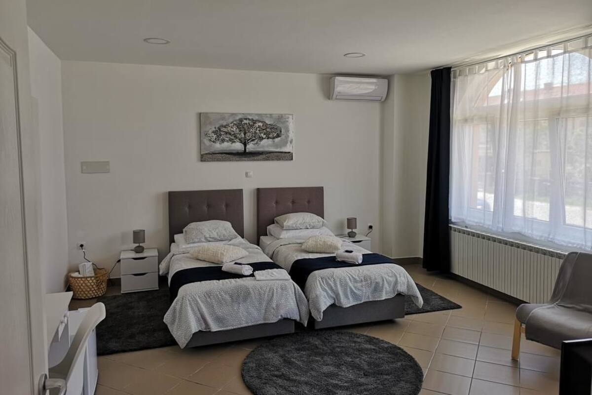 Ein Schlafzimmer mit zwei Betten, einem großen Fenster mit durchsichtigen Vorhängen, einem Heizkörper und einem Bild an der Wand.