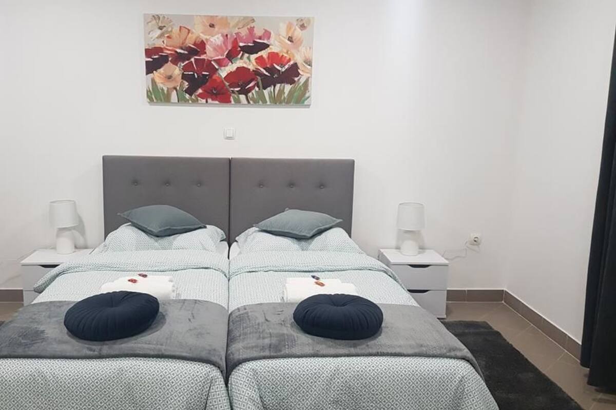 Ein Doppelzimmer mit zwei Betten, jeweils mit grauer Kopfteil und passenden Kissen. Darüber an der Wand hängt ein Blumenbild.