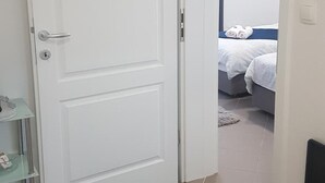 Ein Zimmer mit einer weißen Tür, einem Bett mit weißer Bettwäsche und einem kleinen Regal mit Gegenständen.