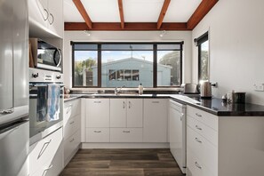 A modern kitchen with white cabinets, stainless steel appliances, and a large window overlooking a blue house. - Classic Kiwi Bach with a modern touch (Dunedin Area)