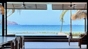 Quarto com uma janela ampla vista para a praia, ventilador de teto e área de refeições com cadeiras e mesa.