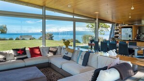 Una sala moderna con un sofá seccional grande, un comedor y una cocina. La habitación tiene ventanas amplias con vista al mar.