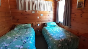 Quarto de cabana com duas camas, paredes de madeira e uma janela com cortinas.