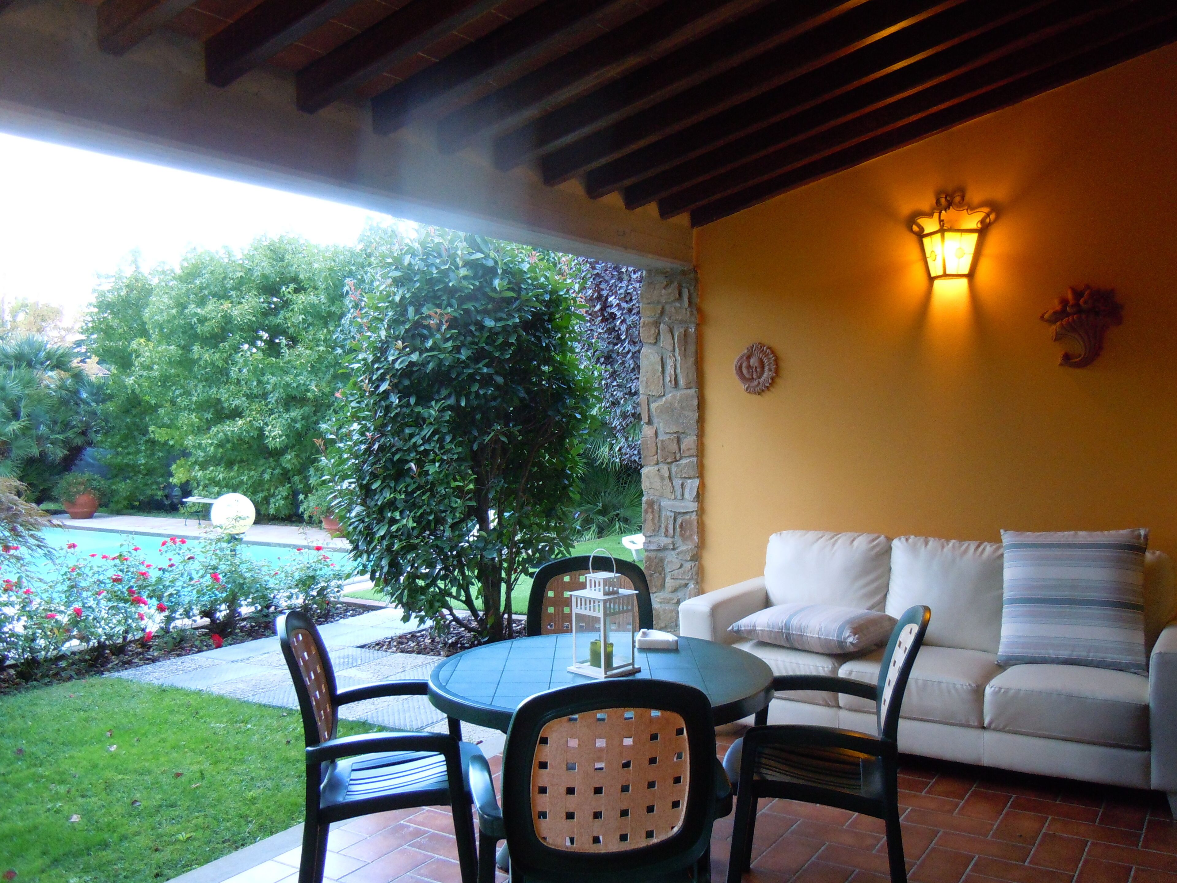 Una terrazza esterna con un tavolo rotondo, sedie e un divano. Si nota un pilastro in pietra, un punto luce a parete e una pianta in vaso.