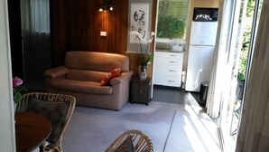 A compact living space with a sofa, dining table, wicker chairs, a small kitchen area, and a view of greenery outside. - Peaceful, sunny, furnished - everything you need. (Johnsonville)