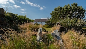 Une maison blanche avec un toit en pierre, entourée de hautes herbes et d’arbres.
