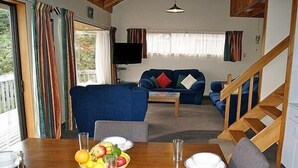 A cozy living room with a blue sofa, a wooden dining table set with plates and a fruit bowl, and a staircase leading to an upper level.