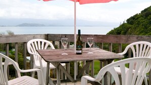 A wooden table with two wine glasses and a bottle, surrounded by white chairs, under a red umbrella, overlooking a lake with hills in the background.