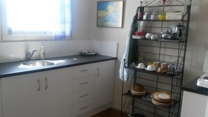 A kitchen with white cabinets, a stainless steel sink, a black countertop, and a metal shelf with dishes and kitchen items. - Historic summer house by the sea (Castlecliff)