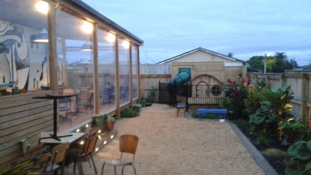 A covered patio area with tables and chairs, a small garden, and a building with a green door. - Historic summer house by the sea (Castlecliff)