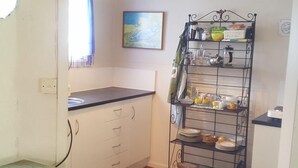 A small kitchen with white cabinets, a black countertop, and a metal shelf unit holding dishes and kitchenware. - Historic summer house by the sea (Castlecliff)
