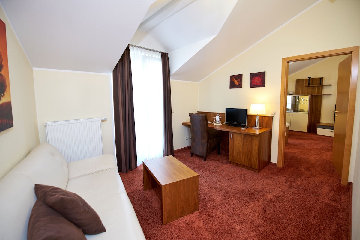 A hotel room with a sofa, a wooden desk, a computer, a lamp, and a view of the kitchen through an open door.